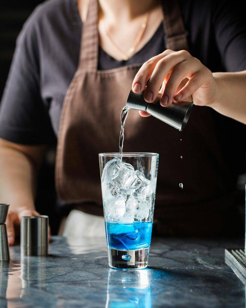 Bartender pouring a clear liquid into a cocktail glass with ice and blue liqueur.